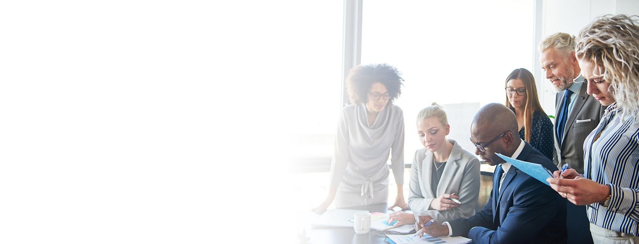 2880x1100 left white gradient of Diverse group of focused businesspeople standing around their manager sitting at an office boardroom table reviewing charts and paperwork