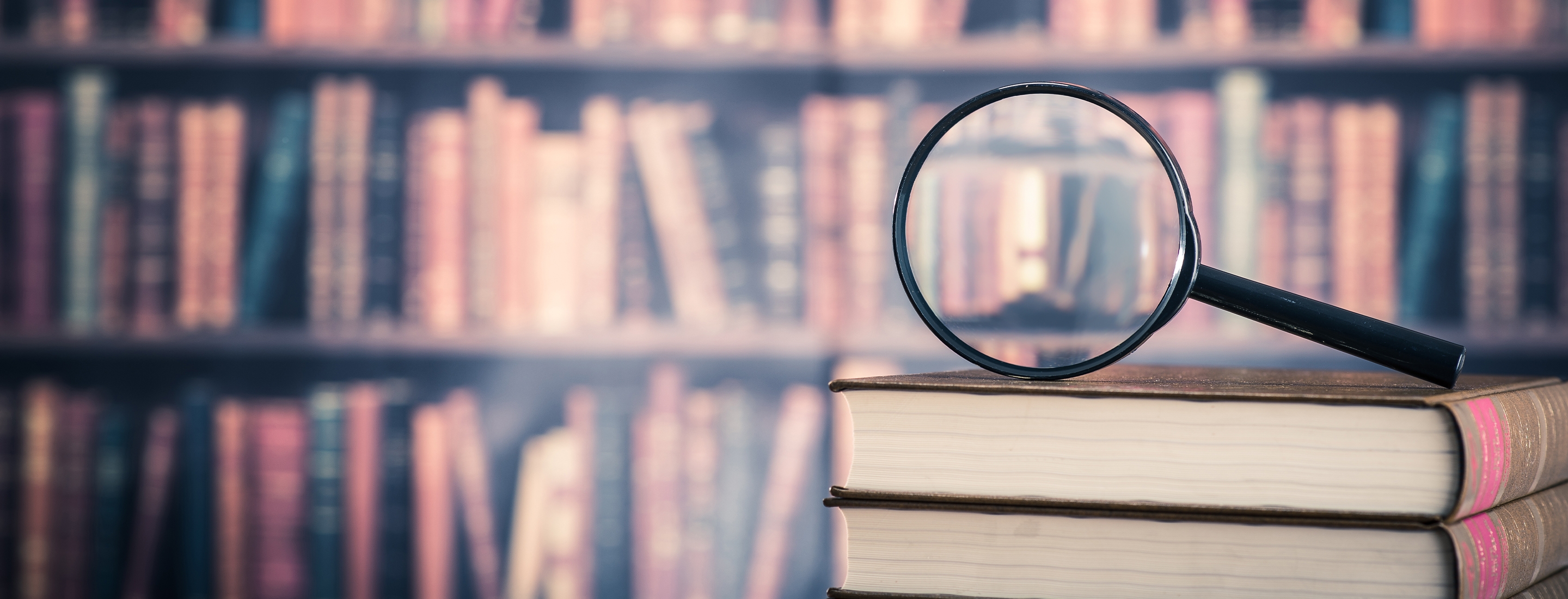 Cropped 2880x1100 of library with books in background and magnifying glass on top of books in the foreground