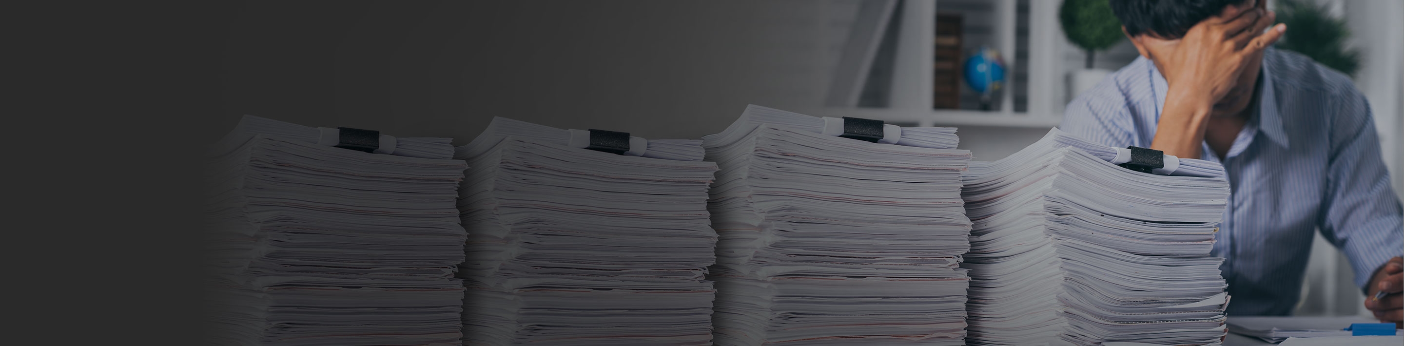 Male with head in his hands at a desk with a large stack of files