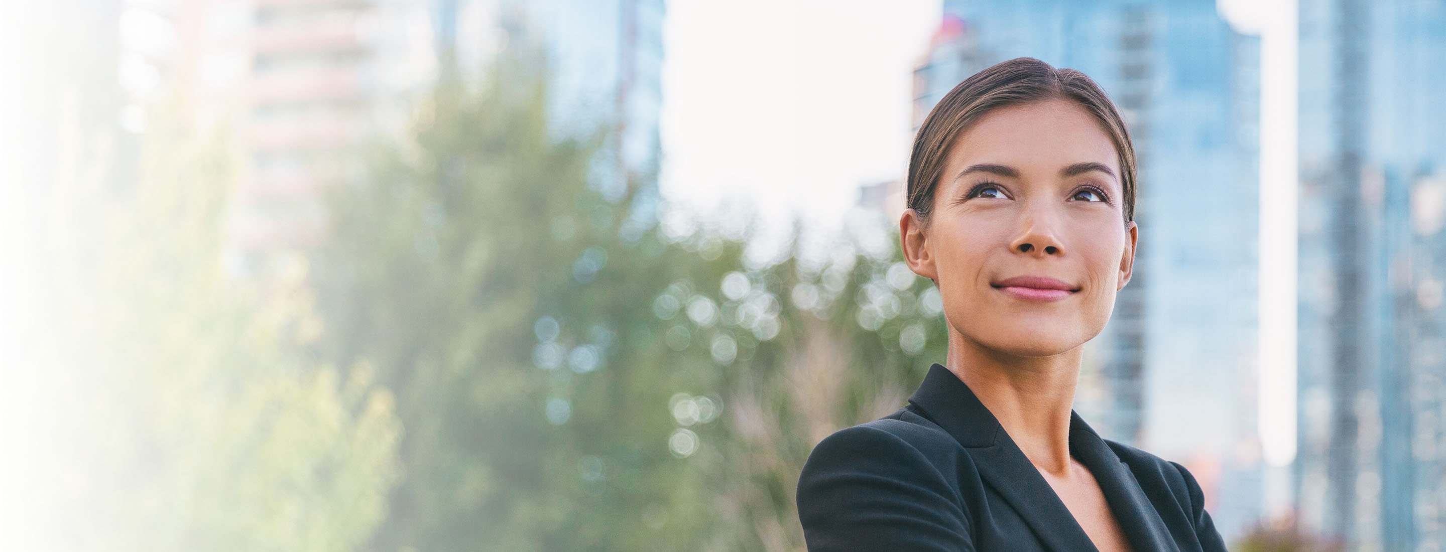 A female lawyer wearing a black blazer in front of a city background looks upward with a small smile on her face.