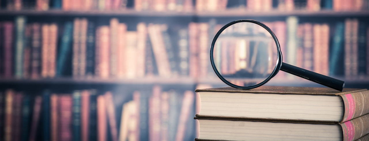 Cropped 2880x1100 of library with books in background and magnifying glass on top of books in the foreground